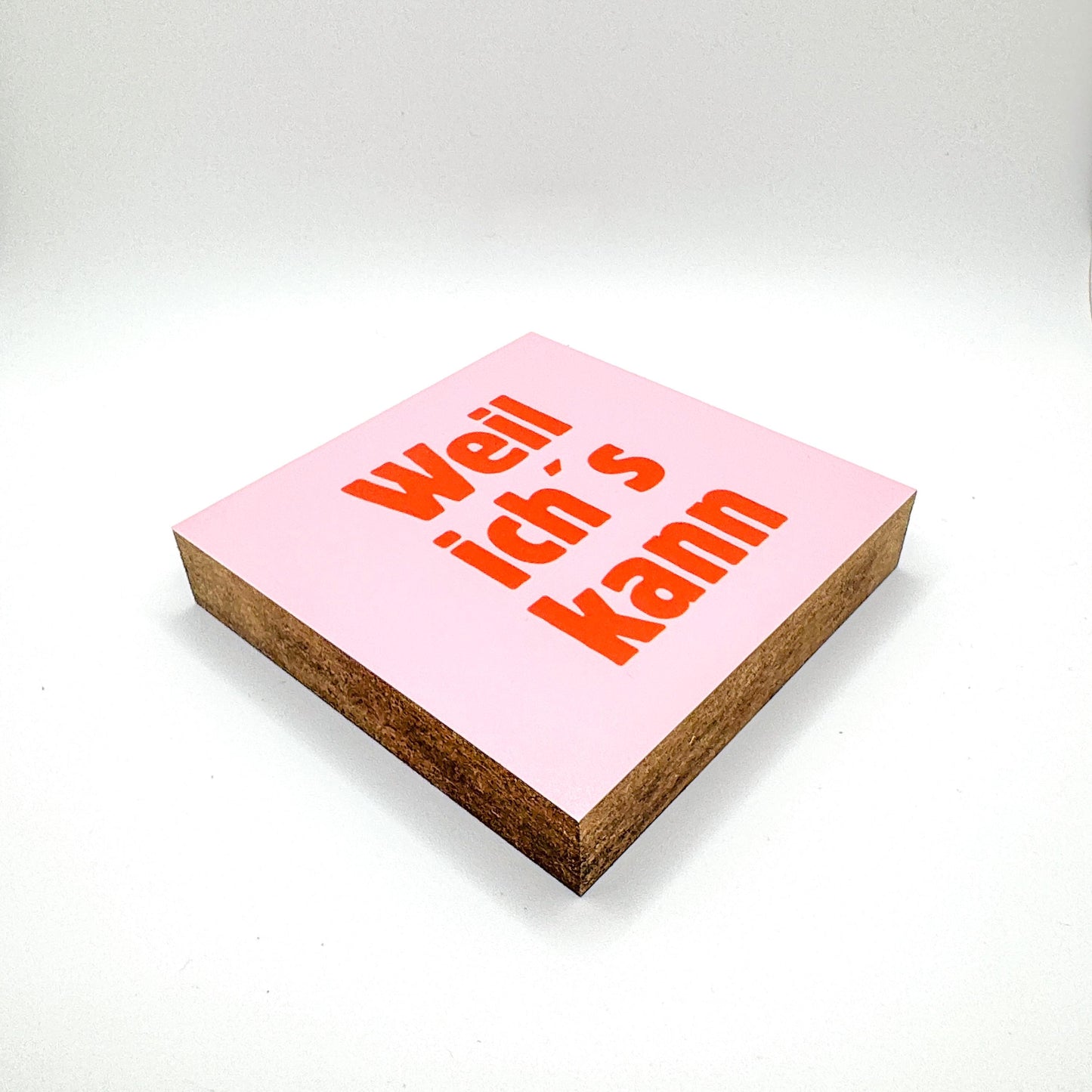 Quadratisches Mini Holzbild mit rosa Hintergrund und orange-rot farbener Schrift „Weil ich’s kann“, schräg liegend aufgenommen, klar lesbar, minimalistische Typografie auf MDF-Holzblock mit Nussbaum Lasur